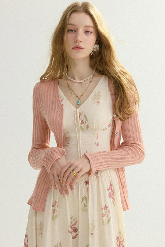 A young woman with long blonde hair models a pink ribbed cardigan over a cream floral dress against a plain light background.