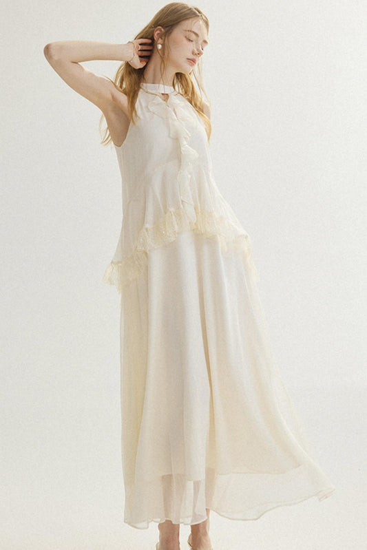 A woman modeling a sleeveless, high-low midi dress in a warm cream or off-white color. The dress features a ruffled tie-neckline and a layered, flowing skirt.
