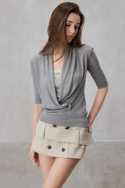 Woman wearing a gray cardigan over a patterned top and beige skirt against a plain background