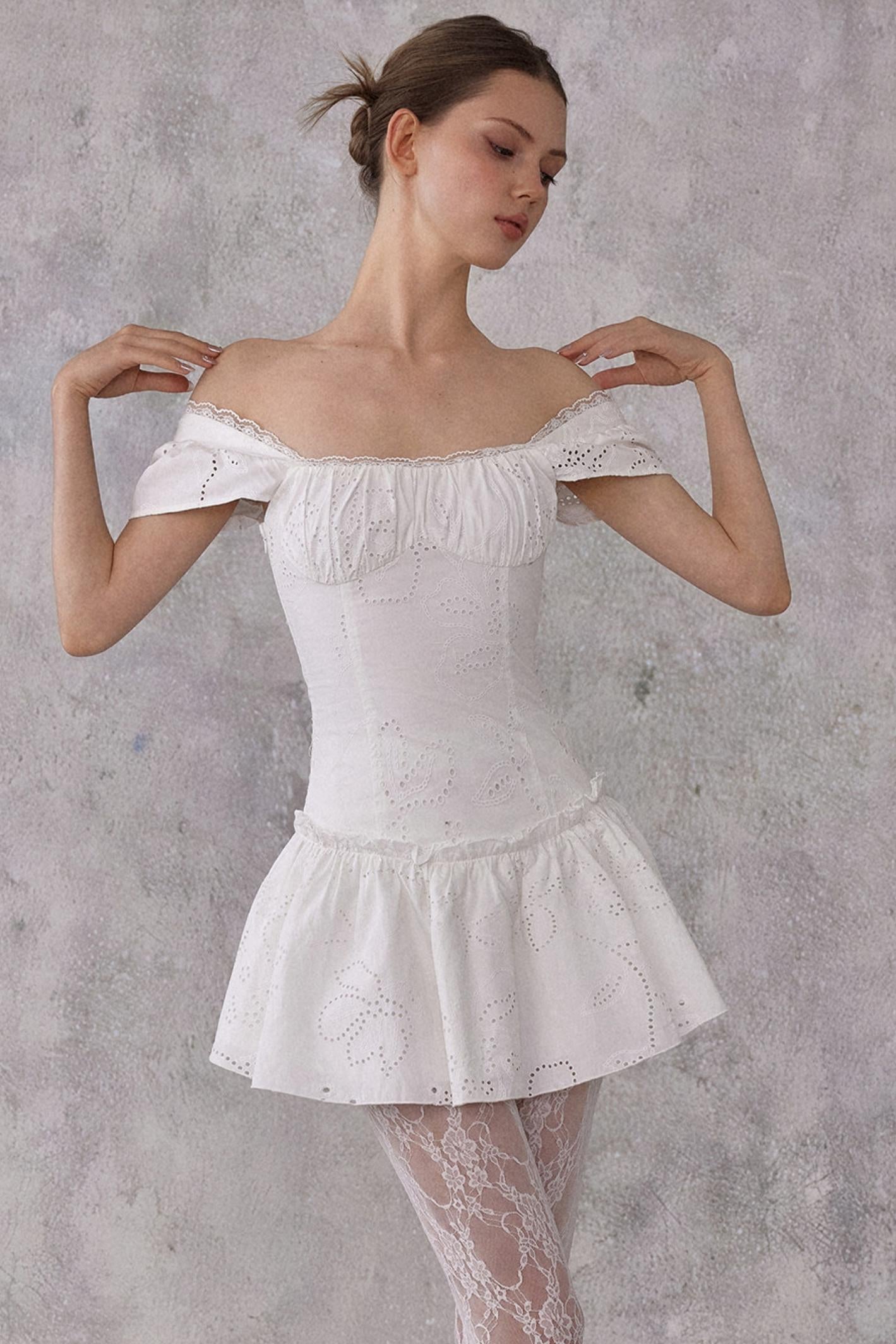 Model styling a romantic white cotton mini dress with eyelet lace details and a smocked pleated waist, worn in a feminine balletcore aesthetic.