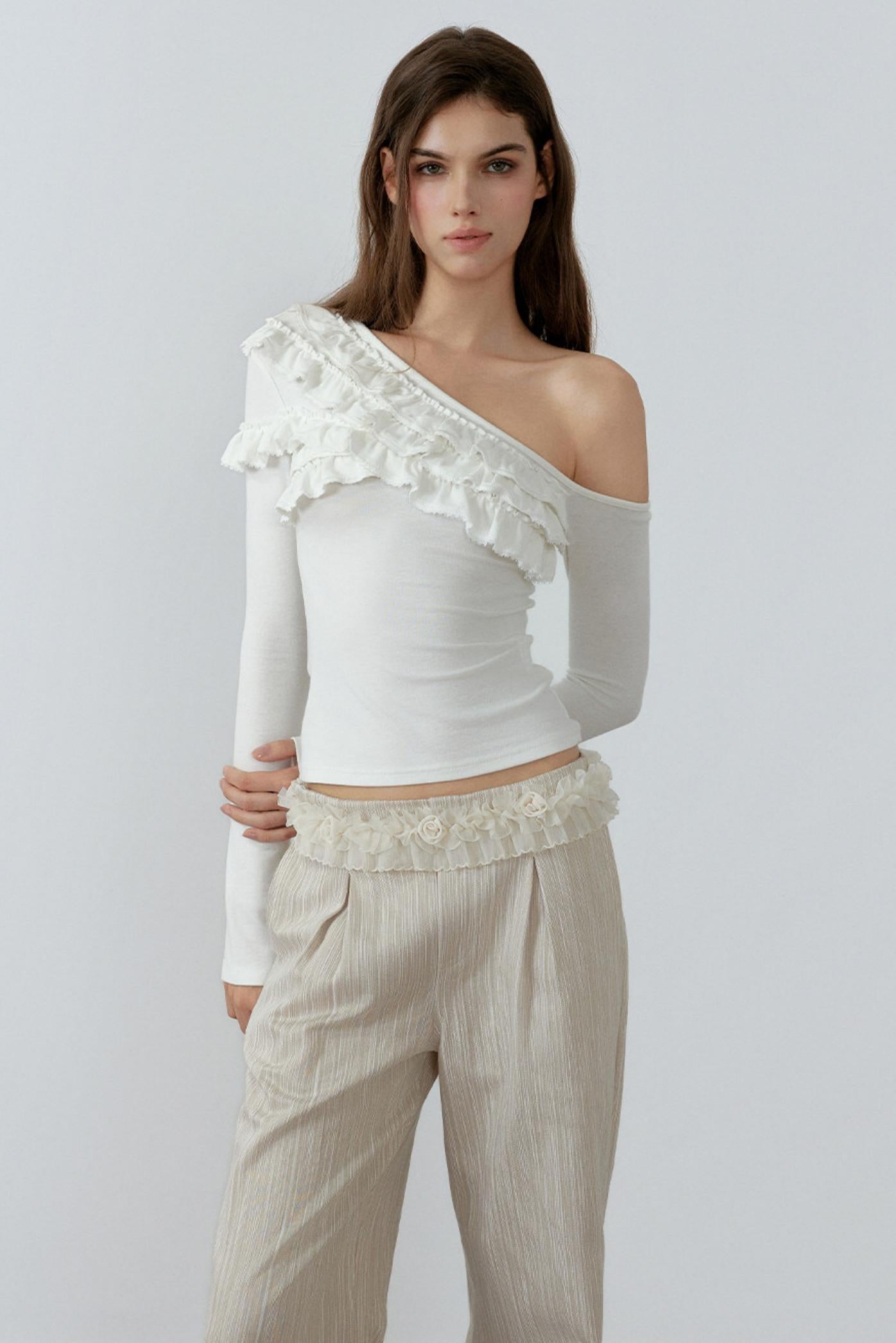 Woman wearing a white one-shoulder top with ruffles and beige pants on a light gray background