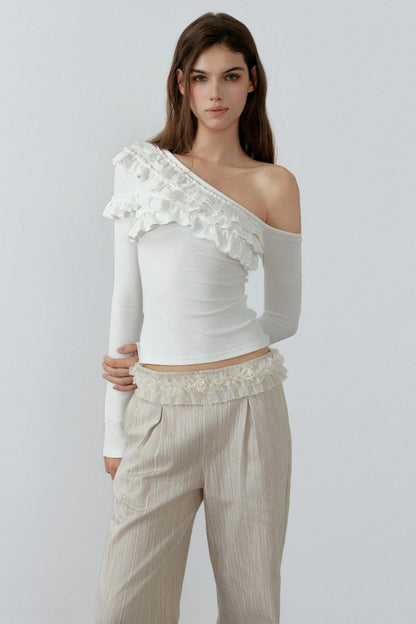 Woman wearing a white one-shoulder top with ruffles and beige pants on a light gray background