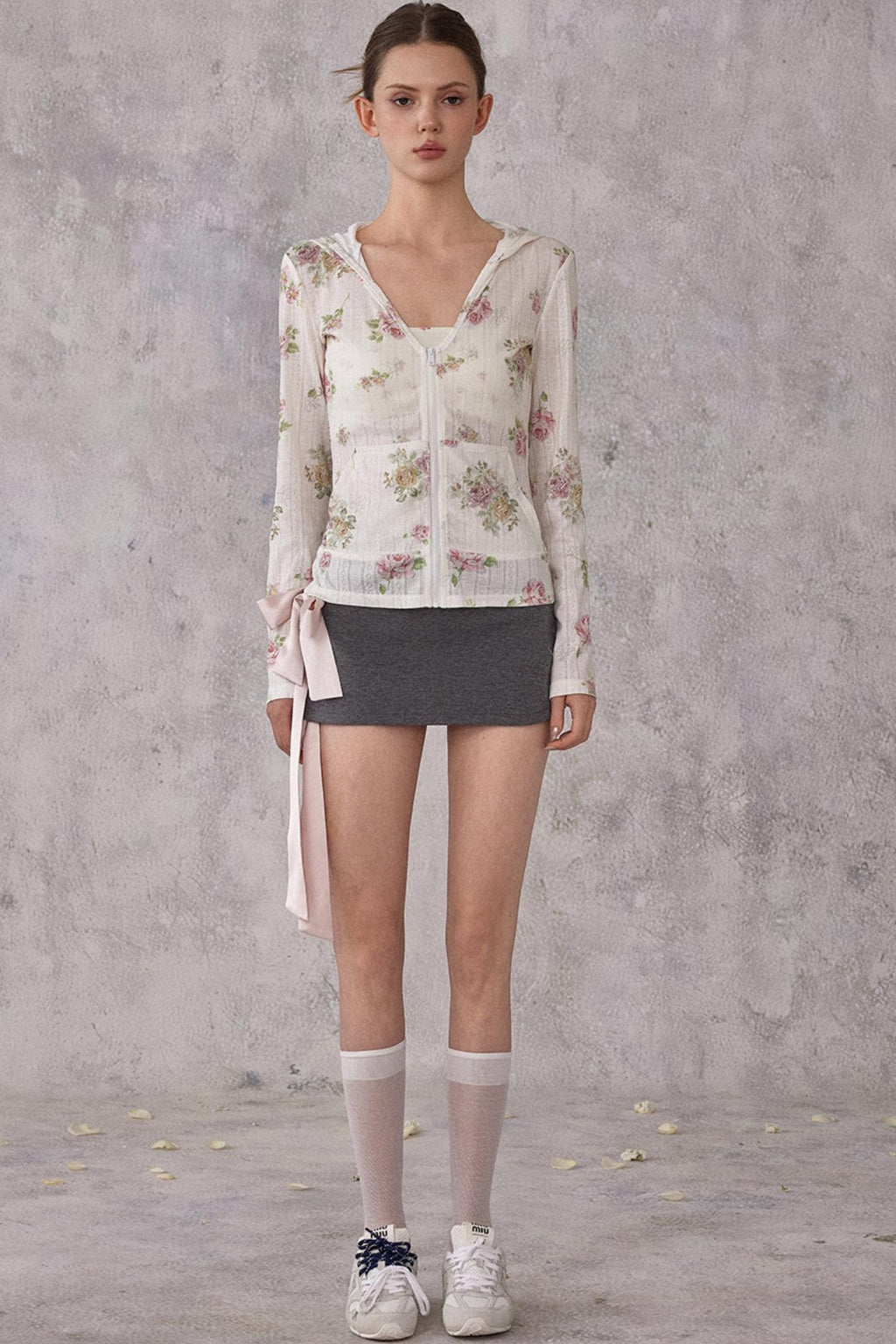 Woman wearing a floral cardigan, black skirt, and white sneakers against a gray concrete wall.