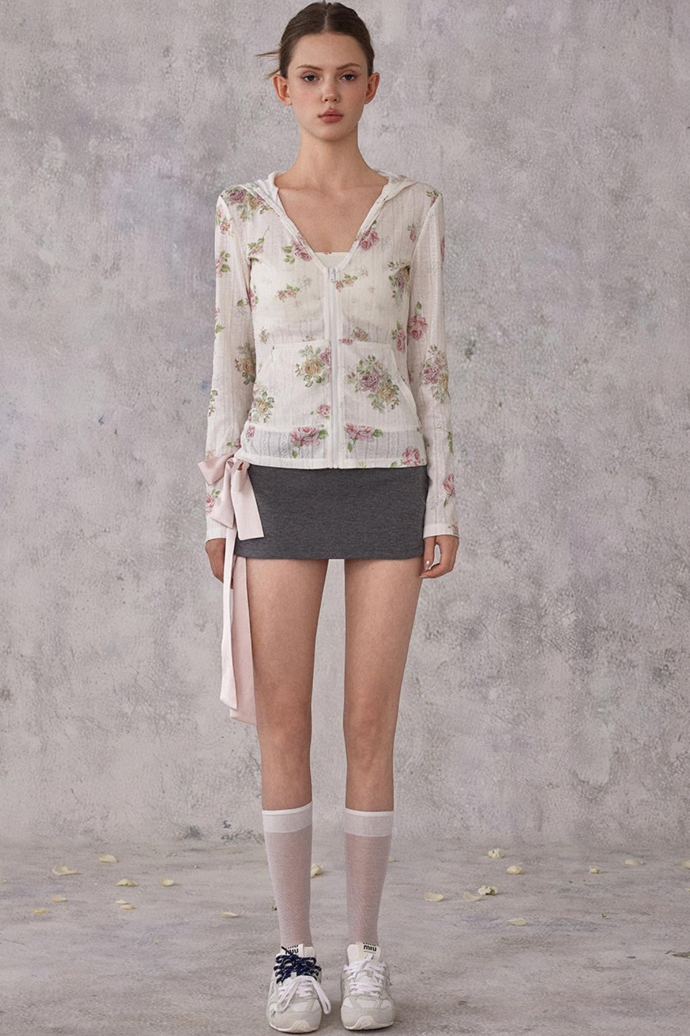Woman wearing a floral cardigan, black skirt, and white sneakers against a gray concrete wall.
