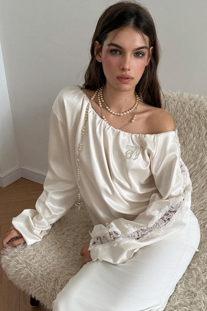 Woman wearing a white satin off-shoulder top with lace details, sitting on a textured surface.