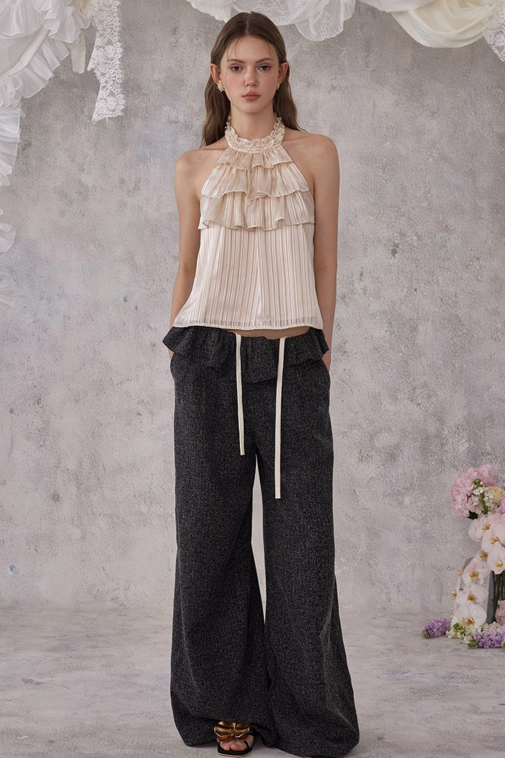 Woman wearing a beige halter top and dark wide-leg pants against a textured wall.