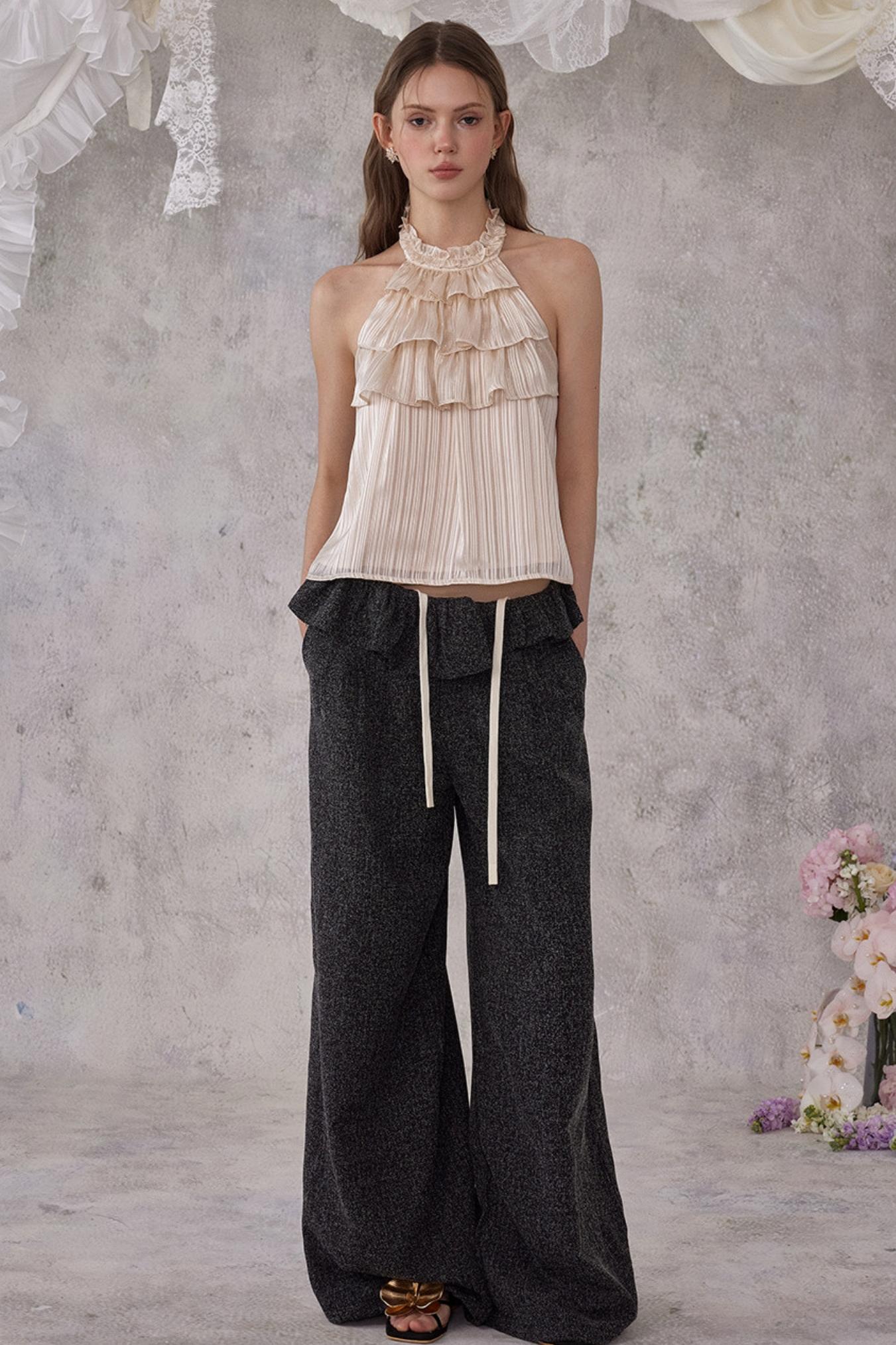 Woman wearing a beige halter top and dark wide-leg pants against a textured wall.