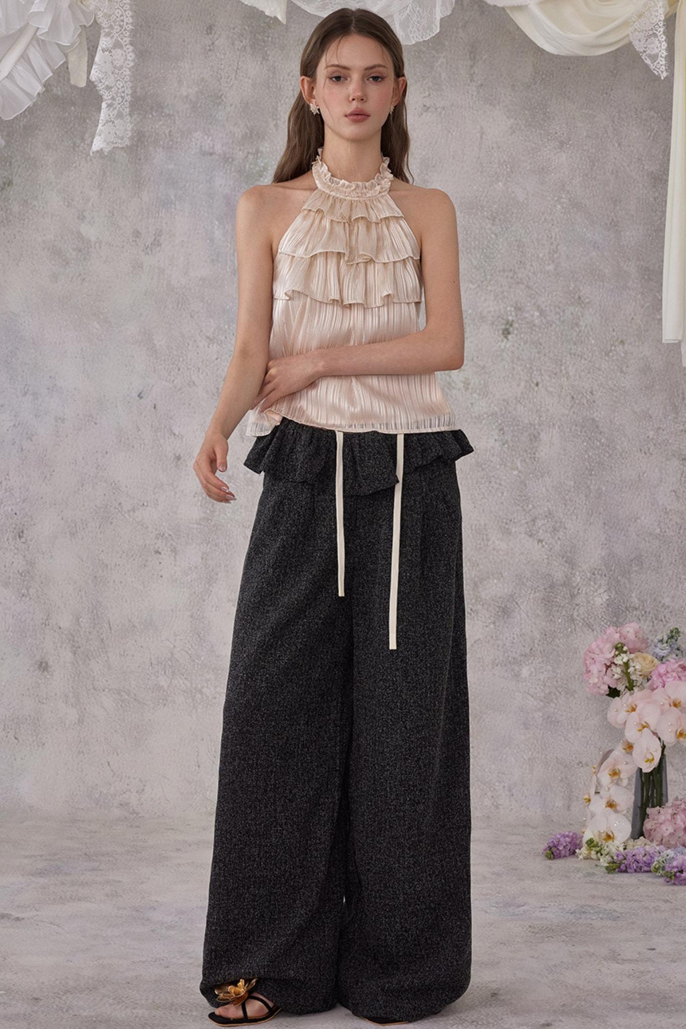 Woman wearing a beige ruffled top and black wide-leg pants against a gray background with flowers.