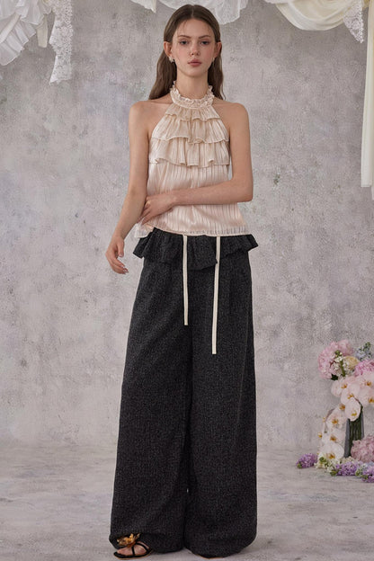 Woman wearing a beige ruffled top and black wide-leg pants against a gray background with flowers.