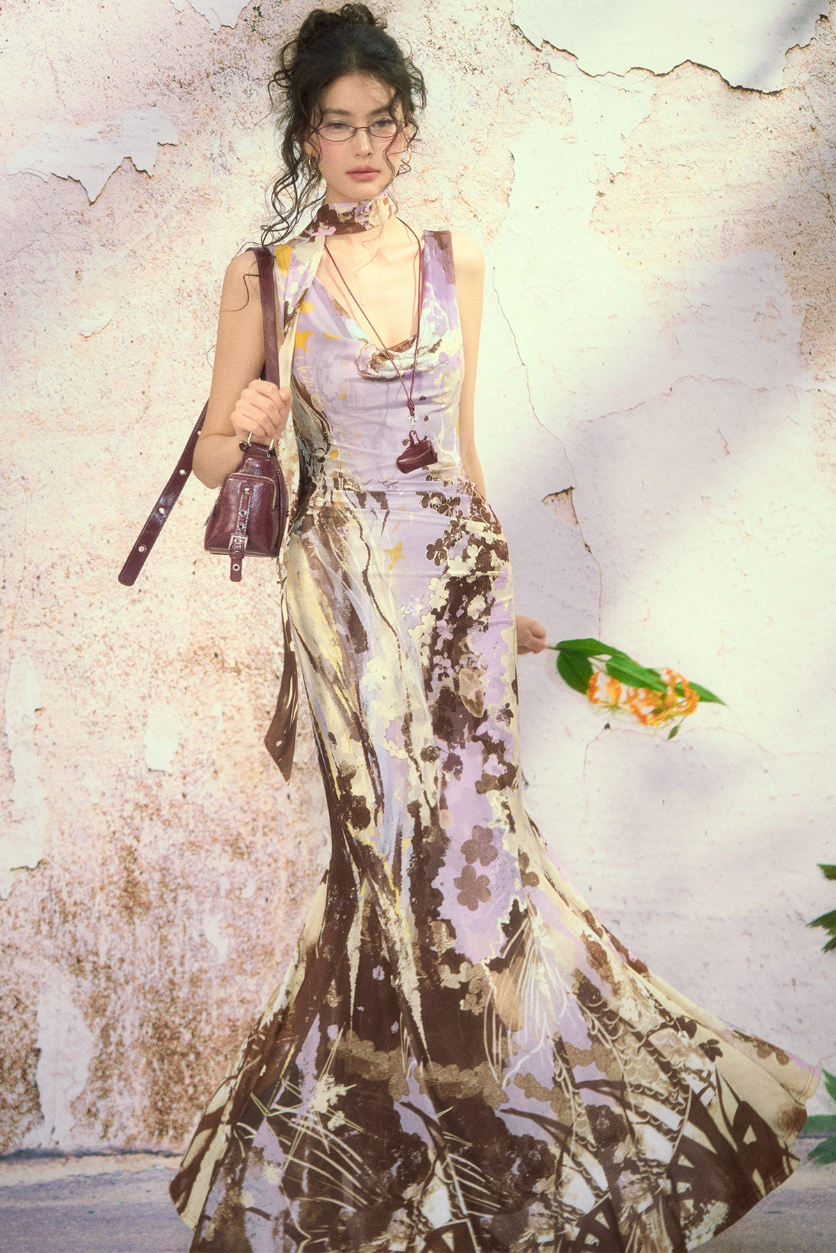 A woman wearing a sleeveless, floor-length halter dress with a lavender, brown, and beige abstract botanical print, walking against a textured, peeling plaster wall while carrying a dark purple handbag.