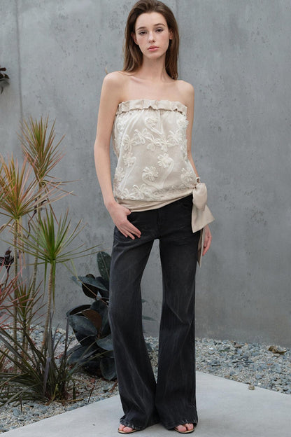 Woman wearing a strapless floral top and black pants against a gray wall.