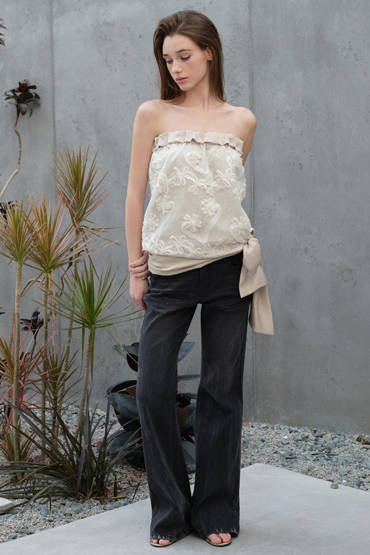 Woman wearing a strapless floral top and black pants standing against a gray wall with plants.