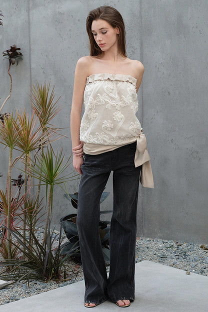 Woman wearing a strapless floral top and black pants standing against a gray wall with plants.