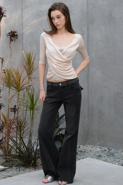 Woman wearing a beige top and black pants standing against a gray wall with plants.