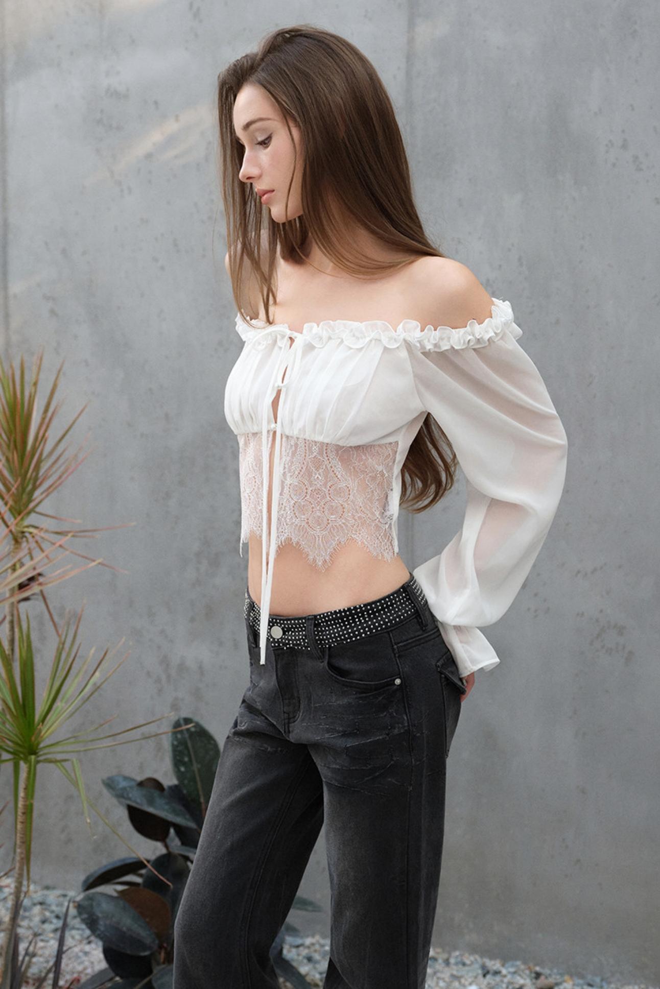 Woman wearing a white off-shoulder top and black pants against a gray wall.