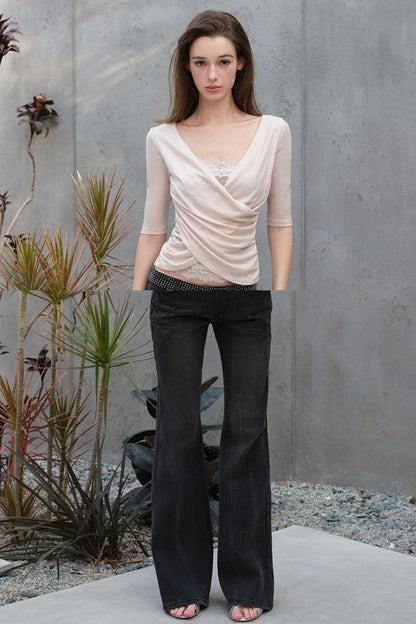 Woman wearing a beige top and black pants standing against a gray wall with plants.
