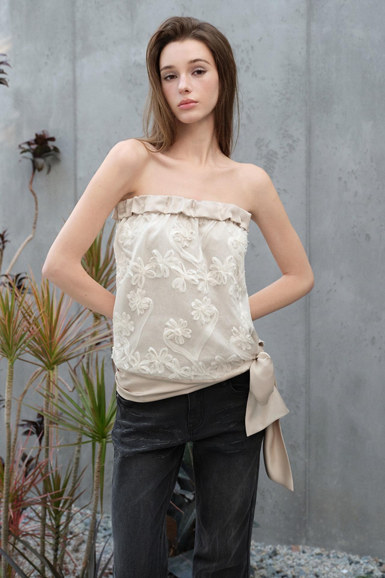 Woman wearing a strapless floral top and dark pants against a gray wall.