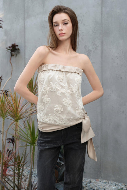 Woman wearing a strapless floral top and dark pants against a gray wall.
