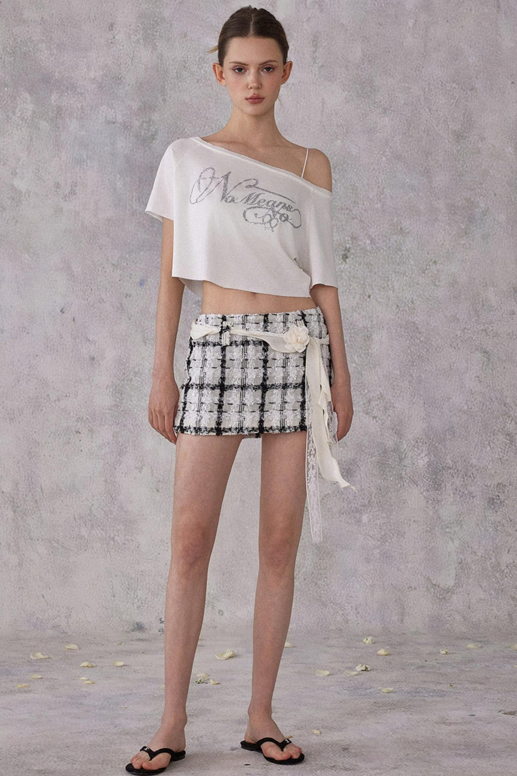 Model wearing a white off-shoulder top and black and white checkered shorts against a gray background