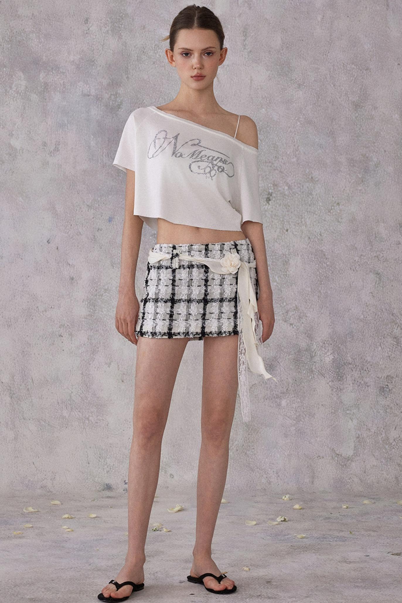 Model wearing a white off-shoulder top and black and white checkered shorts against a gray background