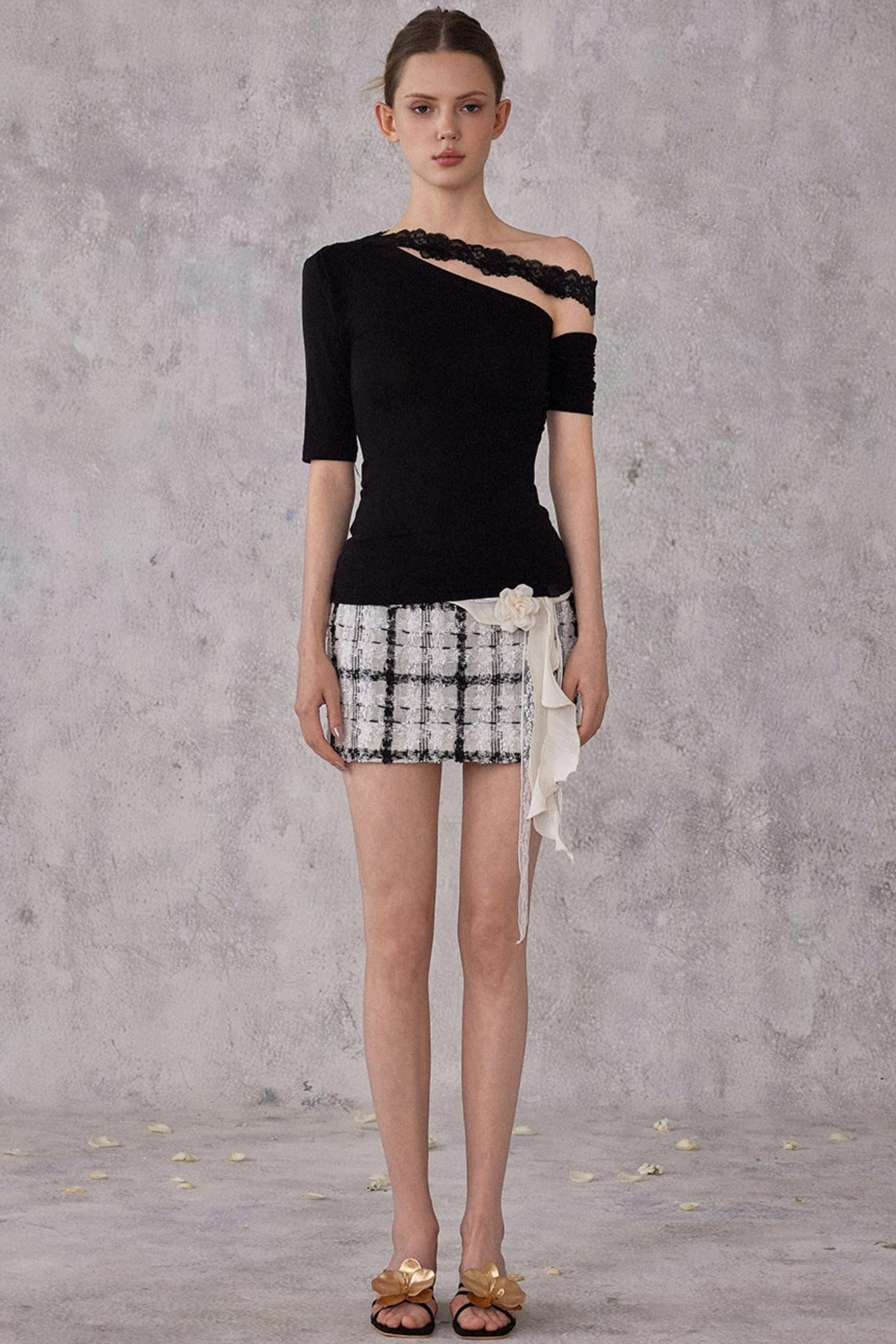Model wearing a black one-shoulder top and plaid shorts against a gray background