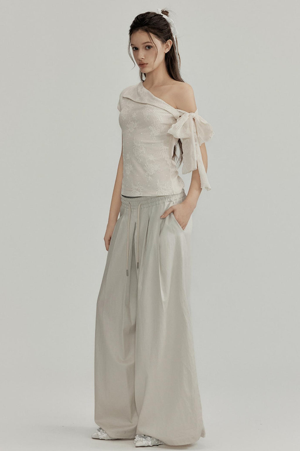 Woman wearing a one-shoulder top with a bow and wide-leg pants on a plain background
