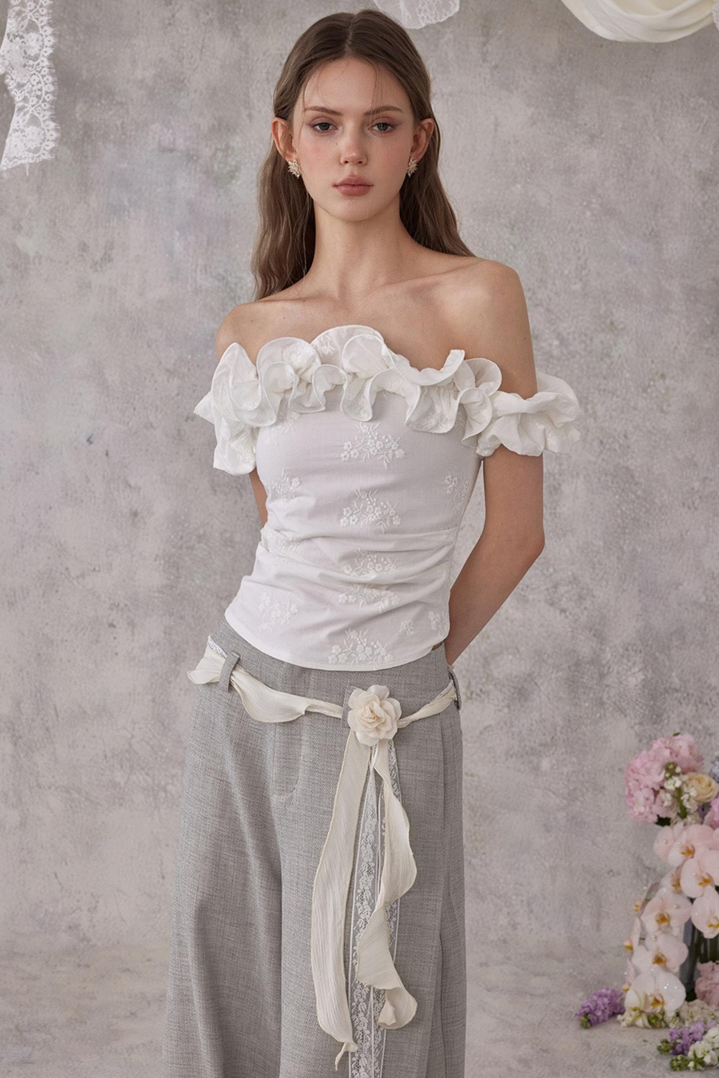 Woman wearing a white off-shoulder top with ruffles and gray pants against a gray background.