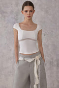 Model wearing a white top with sheer panels and gray pants against a gray background