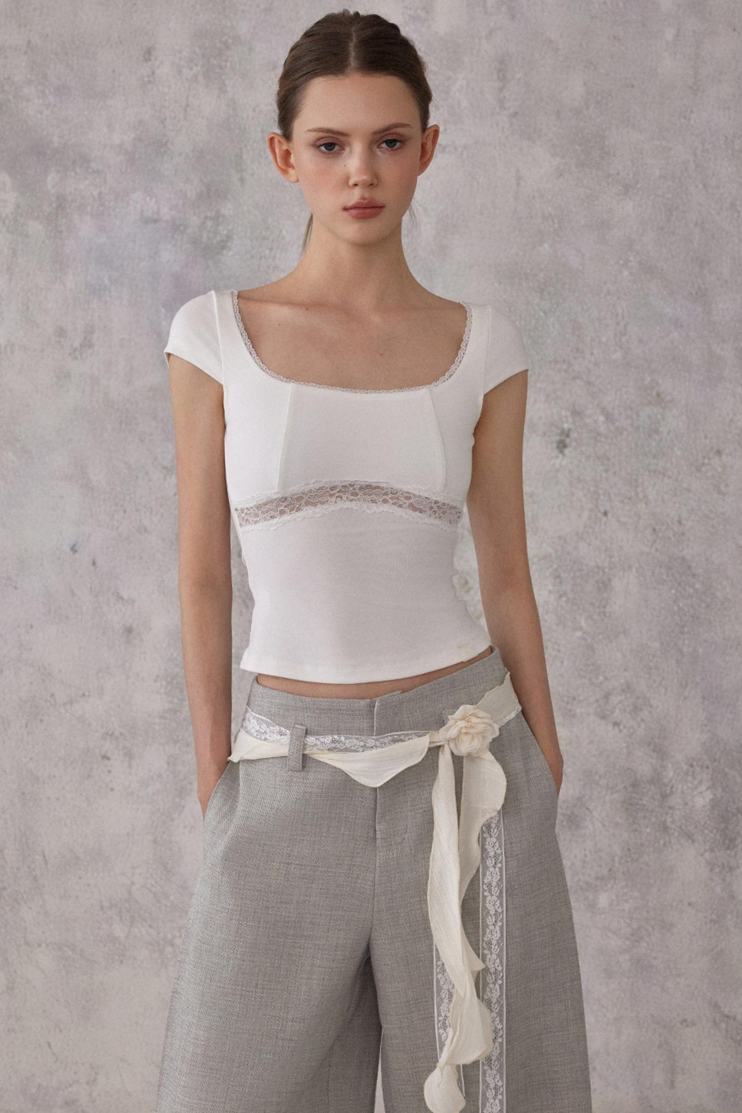 Model wearing a white top with sheer panels and gray pants against a gray background