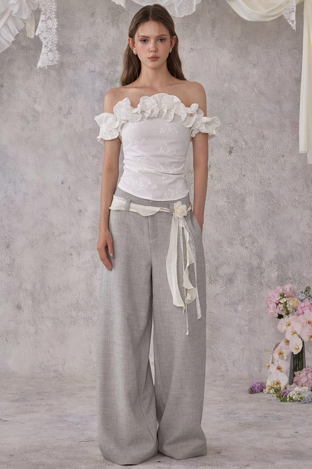 Woman wearing a white off-shoulder top and gray wide-leg pants against a gray background with floral decorations.