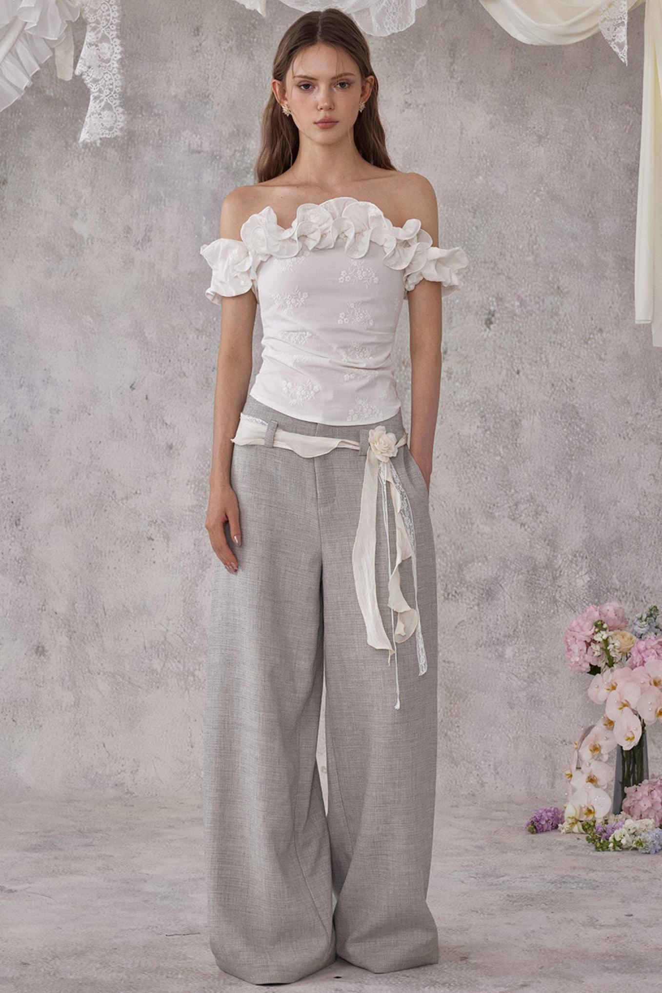 Woman wearing a white off-shoulder top and gray wide-leg pants against a gray background with floral decorations.
