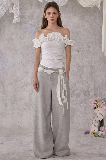 Woman wearing a white off-shoulder top and gray wide-leg pants against a gray background with floral decorations.