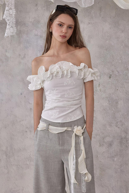 Model styling a romantic white embroidered cotton one-shoulder top with delicate lettuce ruffle edges, paired with grey wide-leg trousers.