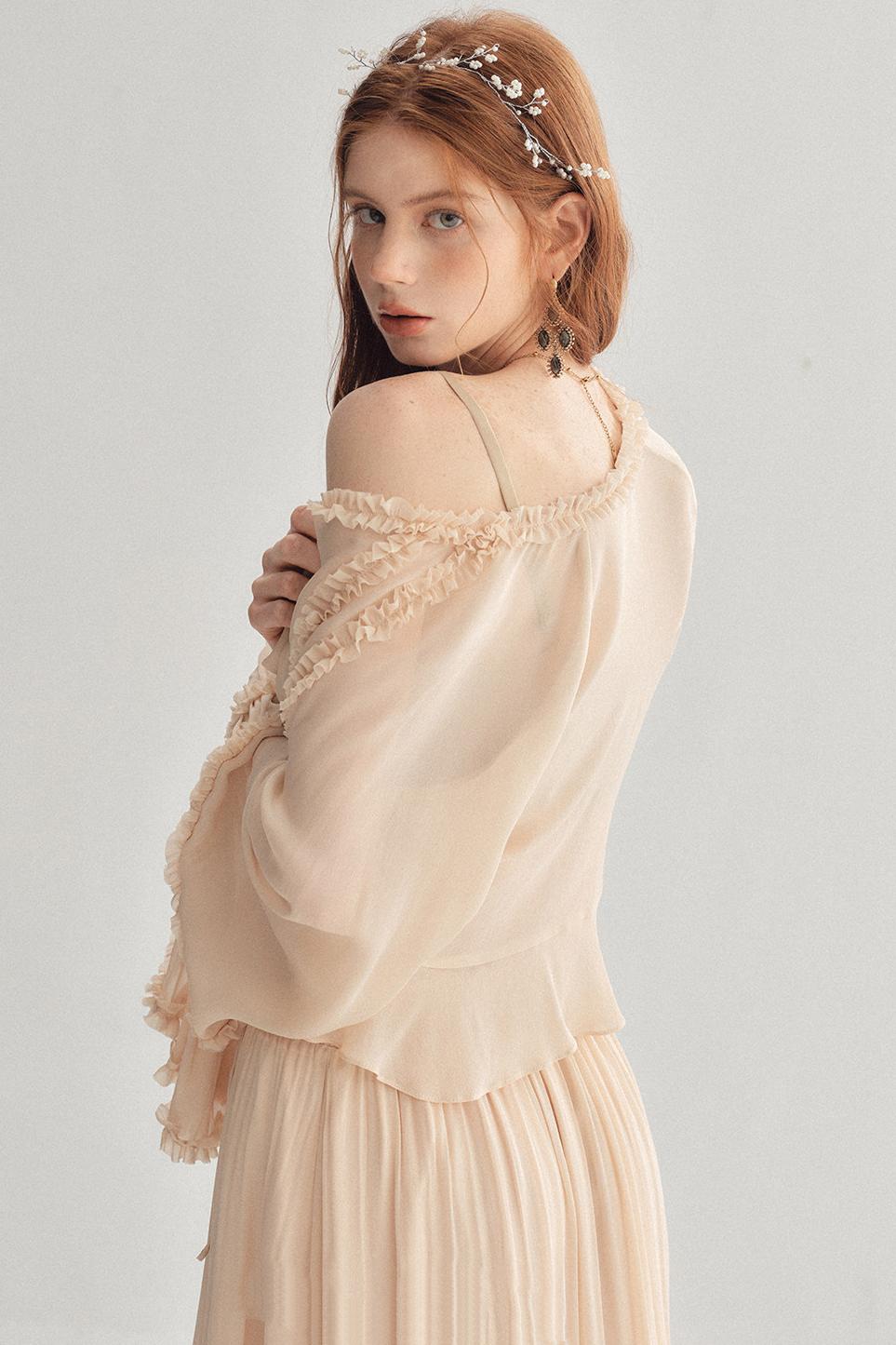 A woman wearing a light beige, off-the-shoulder long-sleeve dress with delicate lace trim and a matching pleated skirt, accessorized with a pearl headband, photographed against a neutral light gray background.
