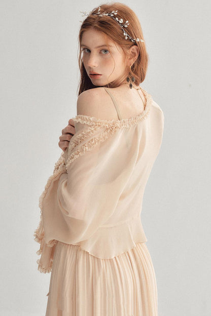 A woman wearing a light beige, off-the-shoulder long-sleeve dress with delicate lace trim and a matching pleated skirt, accessorized with a pearl headband, photographed against a neutral light gray background.