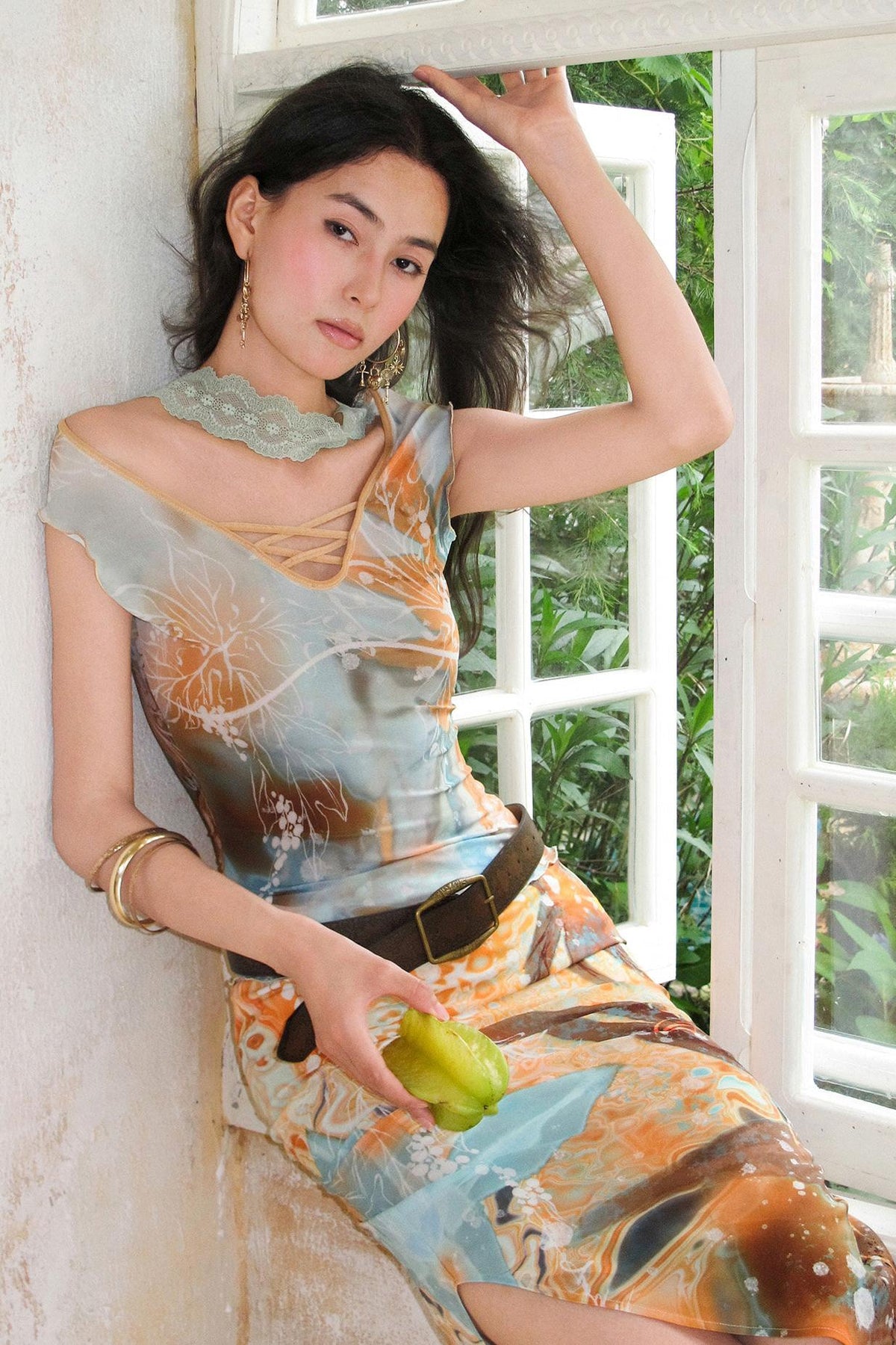 A woman posing in a sleeveless, one-shoulder dress with an abstract orange, blue, and brown print, leaning against a white wall next to a window.