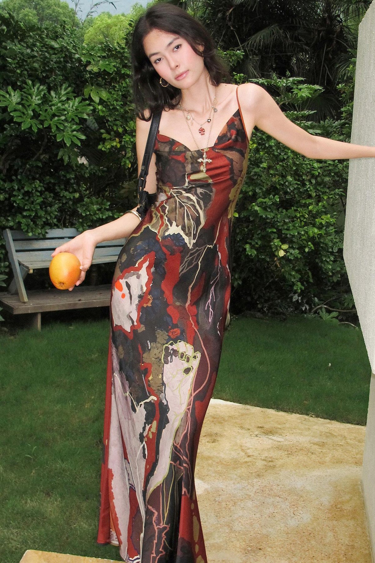 A woman wearing a long, form-fitting spaghetti-strap maxi dress with an abstract painterly print in red, black, brown, and beige, standing outdoors in a garden setting and holding an orange.