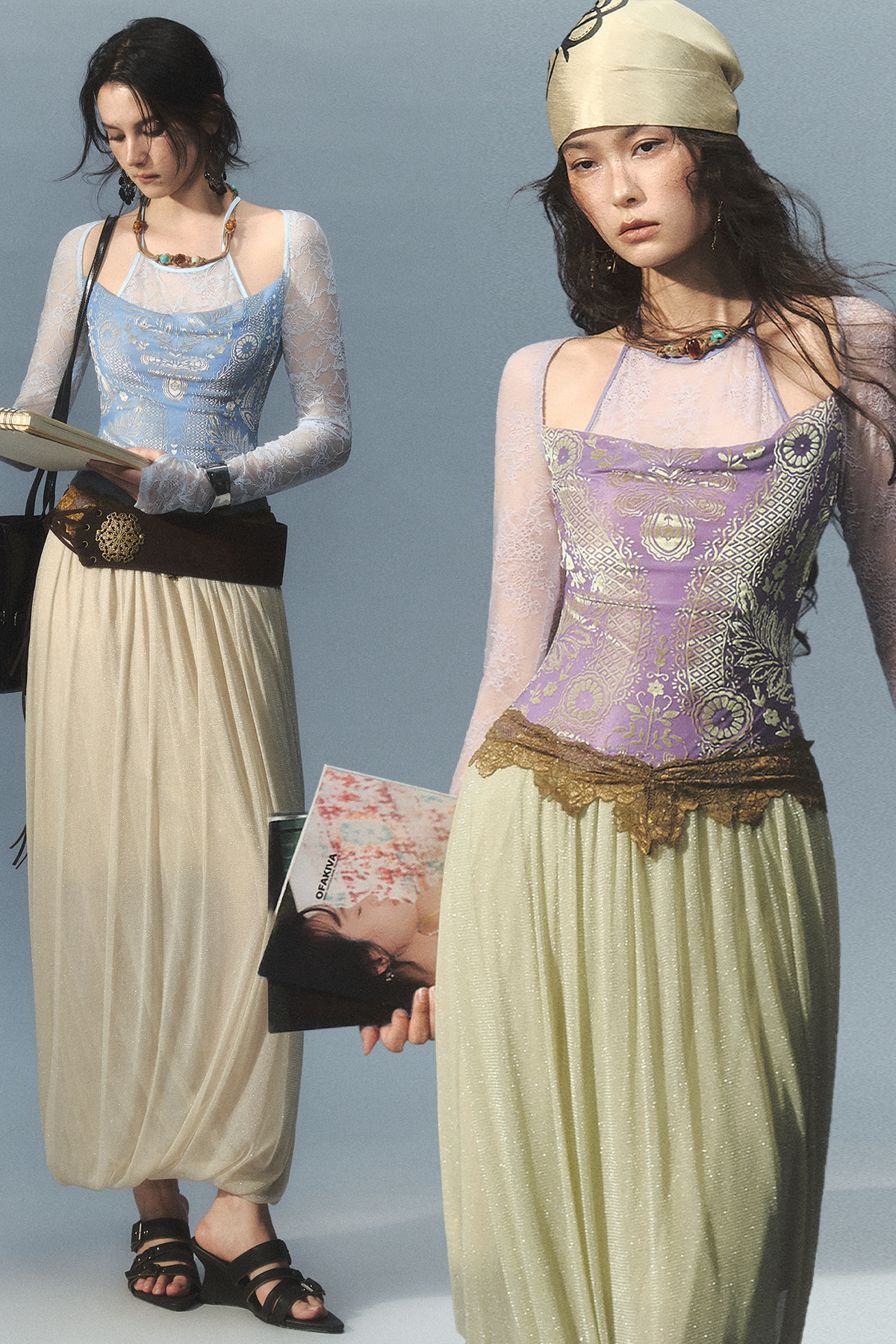 Two models wearing bohemian-style vintage-inspired outfits against a solid light blue-gray studio background. The outfit features a sheer lace long-sleeve top, a patterned camisole, a wide brown belt, and a flowing pleated skirt.
