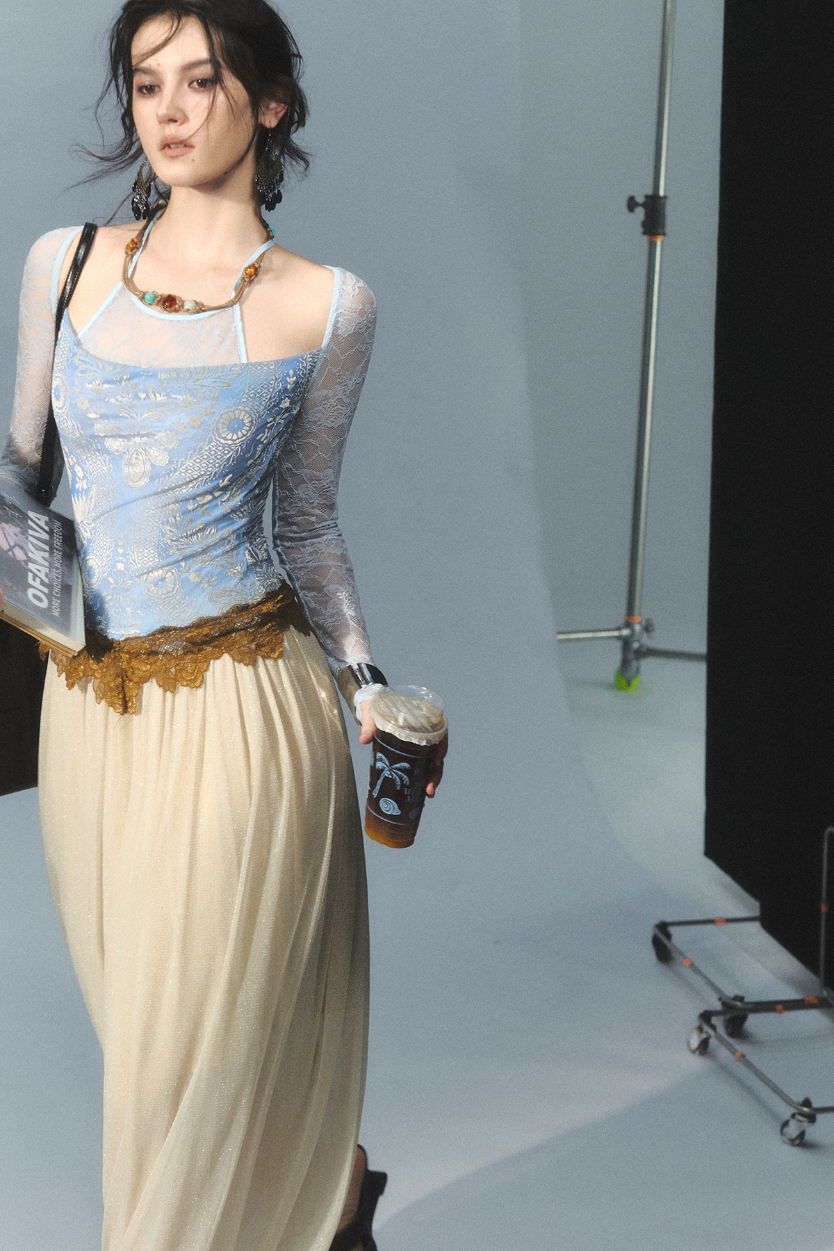 A model walks in a studio wearing a light blue lace-trimmed camisole top with a beige maxi skirt and holding a tiki-style coffee cup.