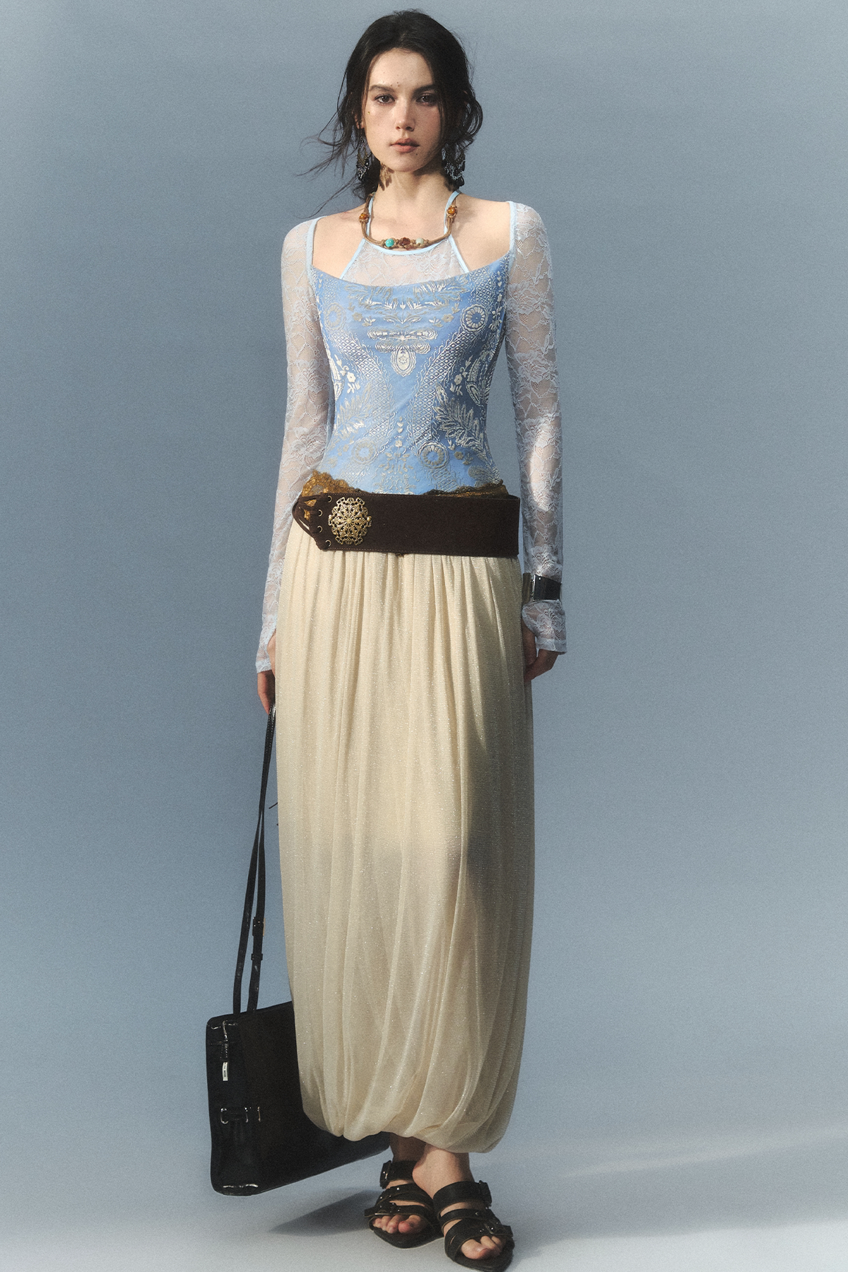 A model stands against a light blue-gray studio background wearing a long-sleeve lace-trimmed top with a light blue embroidered bodice, a cream maxi skirt, a dark brown belt with a decorative gold medallion, and black sandals. She carries a black shoulder bag.