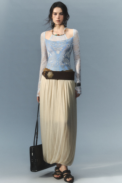A model stands against a light blue-gray studio background wearing a long-sleeve lace-trimmed top with a light blue embroidered bodice, a cream maxi skirt, a dark brown belt with a decorative gold medallion, and black sandals. She carries a black shoulder bag.