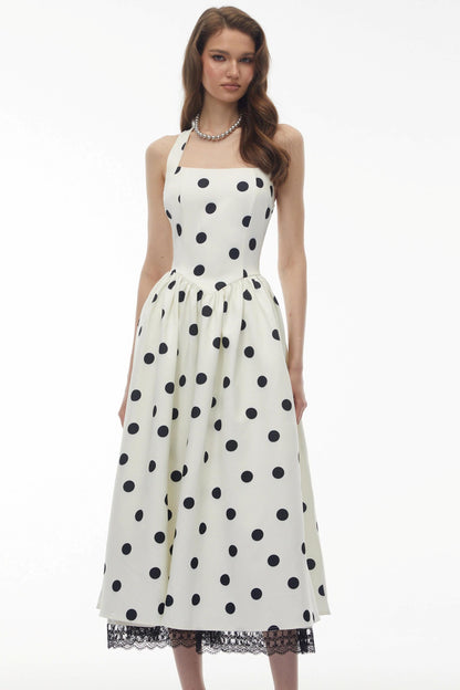 A woman modeling a sleeveless midi dress with a black polka dot pattern on an off-white background, featuring a gathered waist and a lace-trimmed hem, set against a plain white studio background.