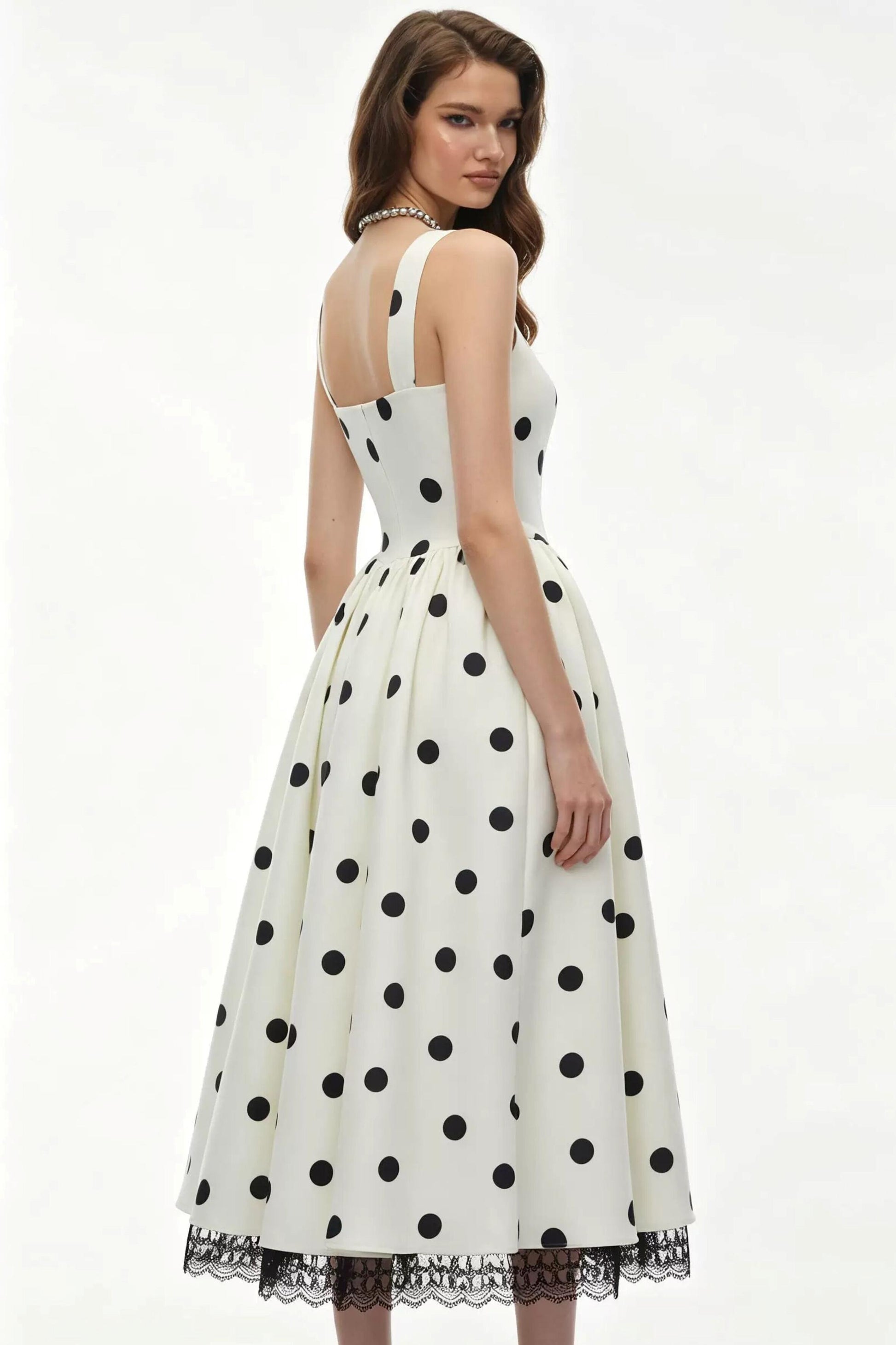 A woman modeling a sleeveless white midi dress with black polka dots and a black lace hem, shown from the back against a plain white studio background.