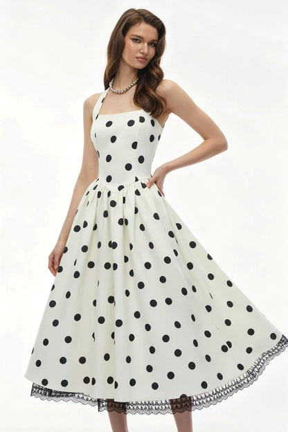 A woman modeling a sleeveless white dress with large black polka dots and a flared skirt against a plain white studio background.