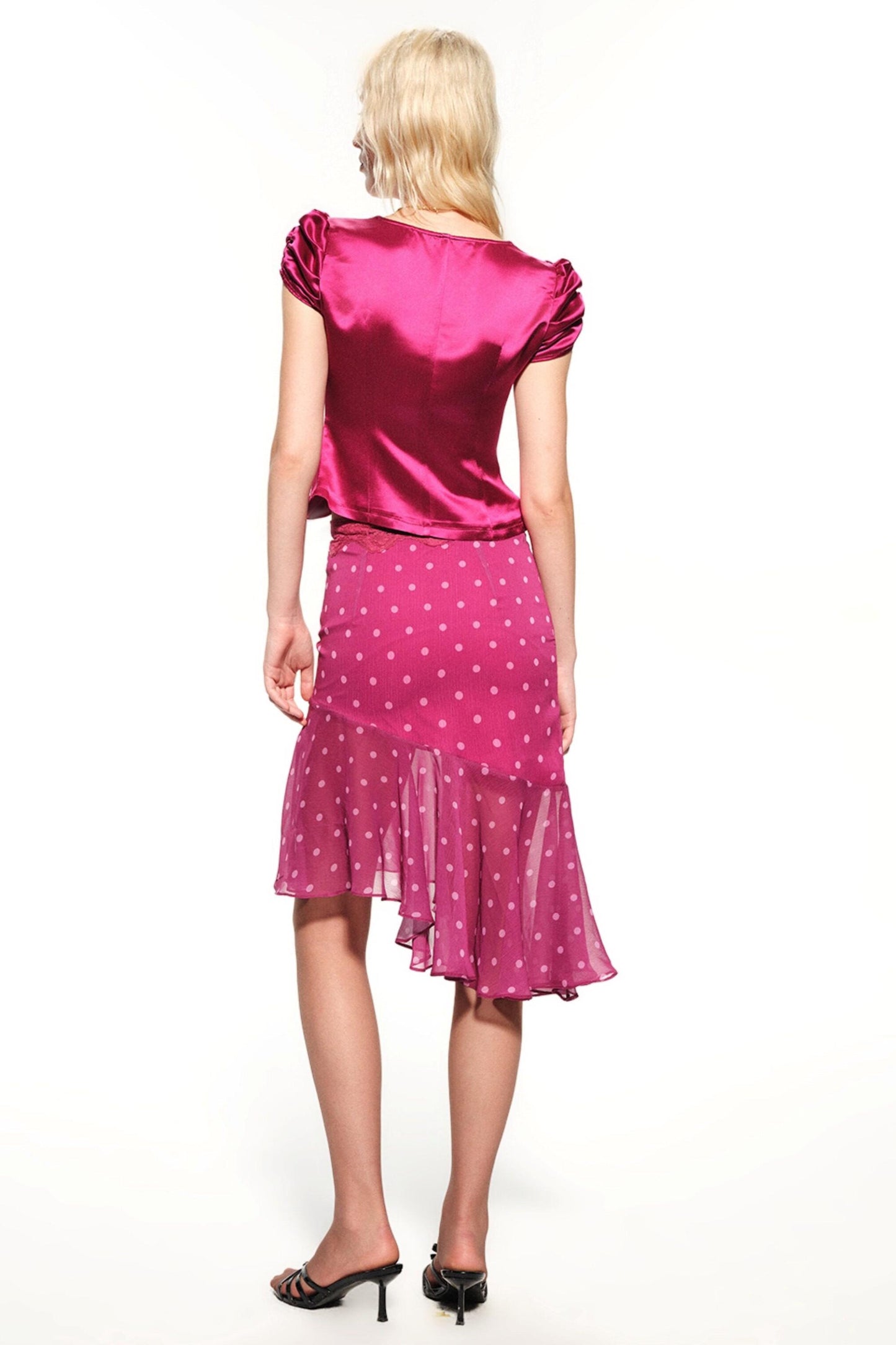 A woman shown from the back wearing a shiny magenta pink satin top with short puff sleeves and a matching pink polka-dot midi skirt with a ruffled hem, paired with black high heels against a white studio background.