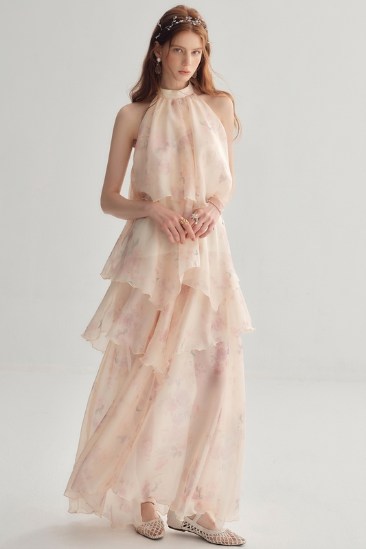 A woman models a sleeveless, high-neck, tiered maxi dress in a very light blush pink with a soft floral print. The dress features a halter-style neckline, a ruffled waist, and a flowing skirt.