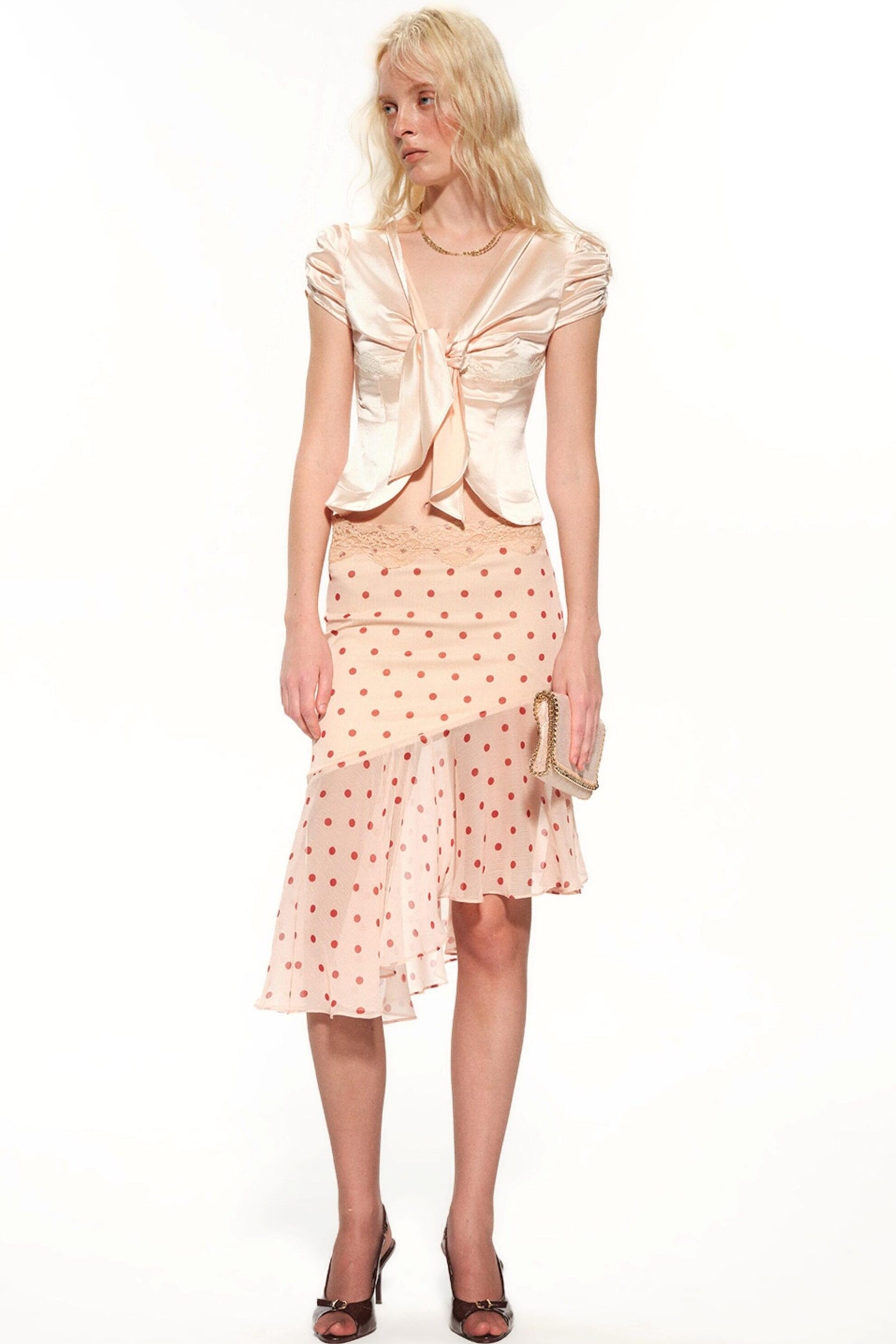 A blonde model wearing a light peach satin top with a large front bow and a matching peach polka-dot skirt, holding a small metallic clutch against a plain white background.