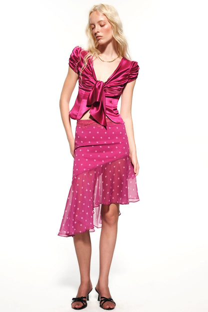 A blonde model wearing a two-piece magenta pink set consisting of a satin-like top with a large front bow and a matching sheer polka-dot skirt, paired with black sandals, on a white studio background.