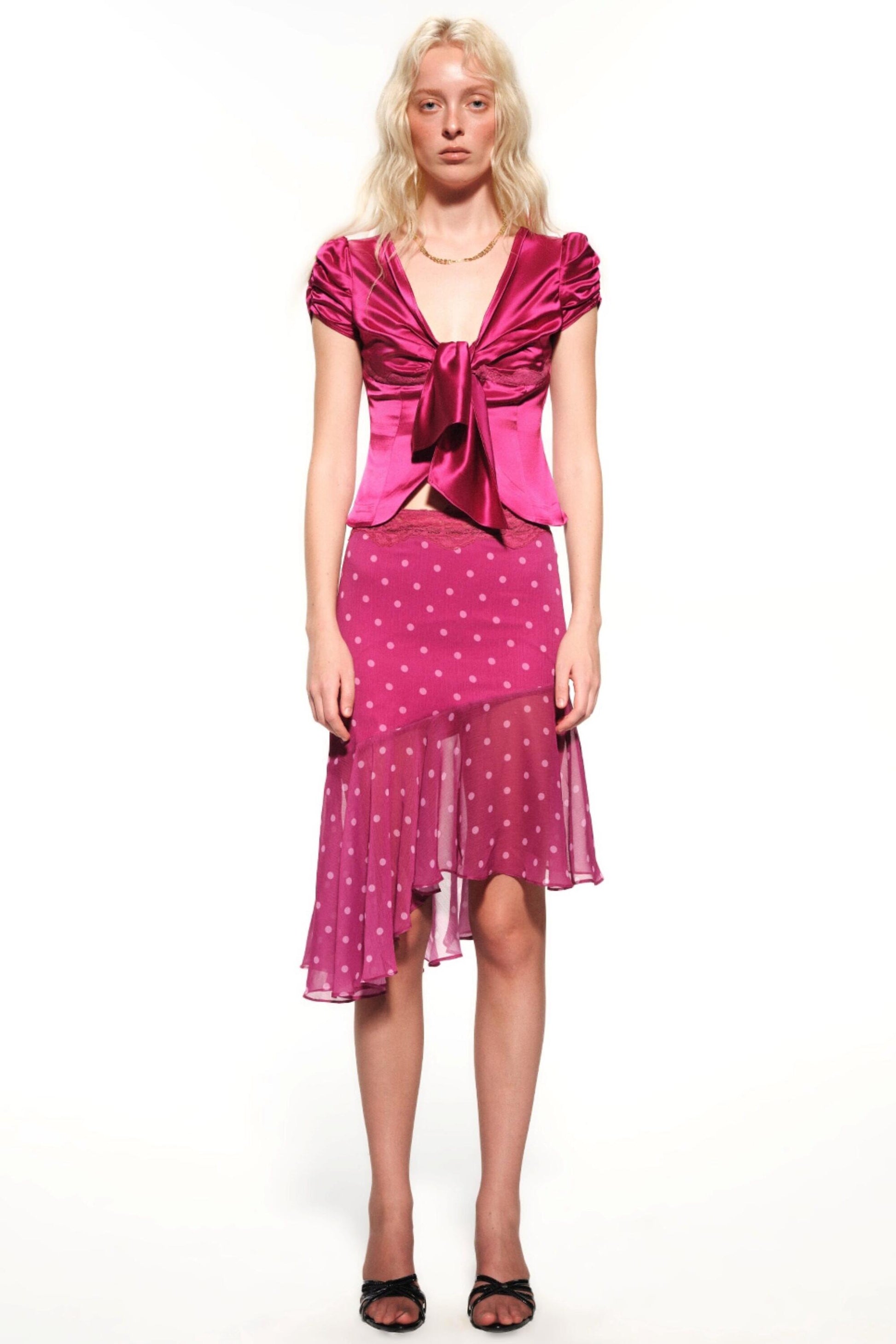 A blonde model stands against a white studio background wearing a bright pink two-piece outfit consisting of a satin-like ruffled top with a large front bow and a matching sheer polka-dot skirt.