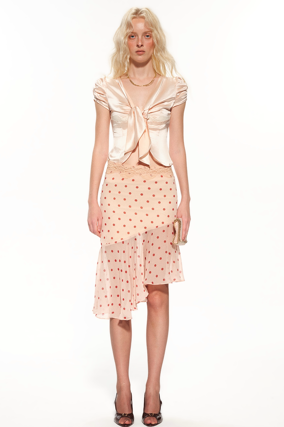 A model stands against a white studio background wearing a light peach satin top with a large front bow and a matching light peach polka-dot skirt with a lace overlay.